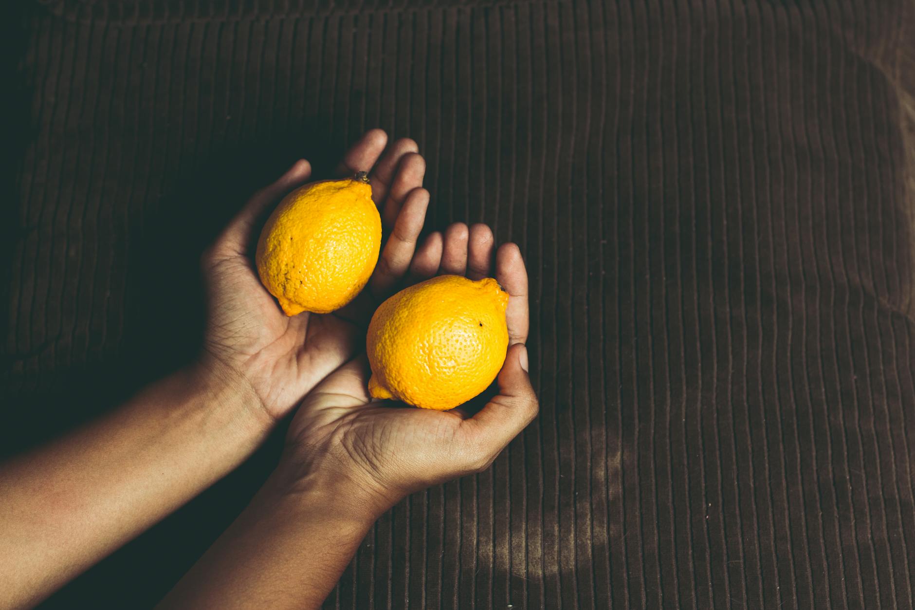 Close-up of two fresh lemons held in cupped hands on a brown surface, symbolizing freshness.
