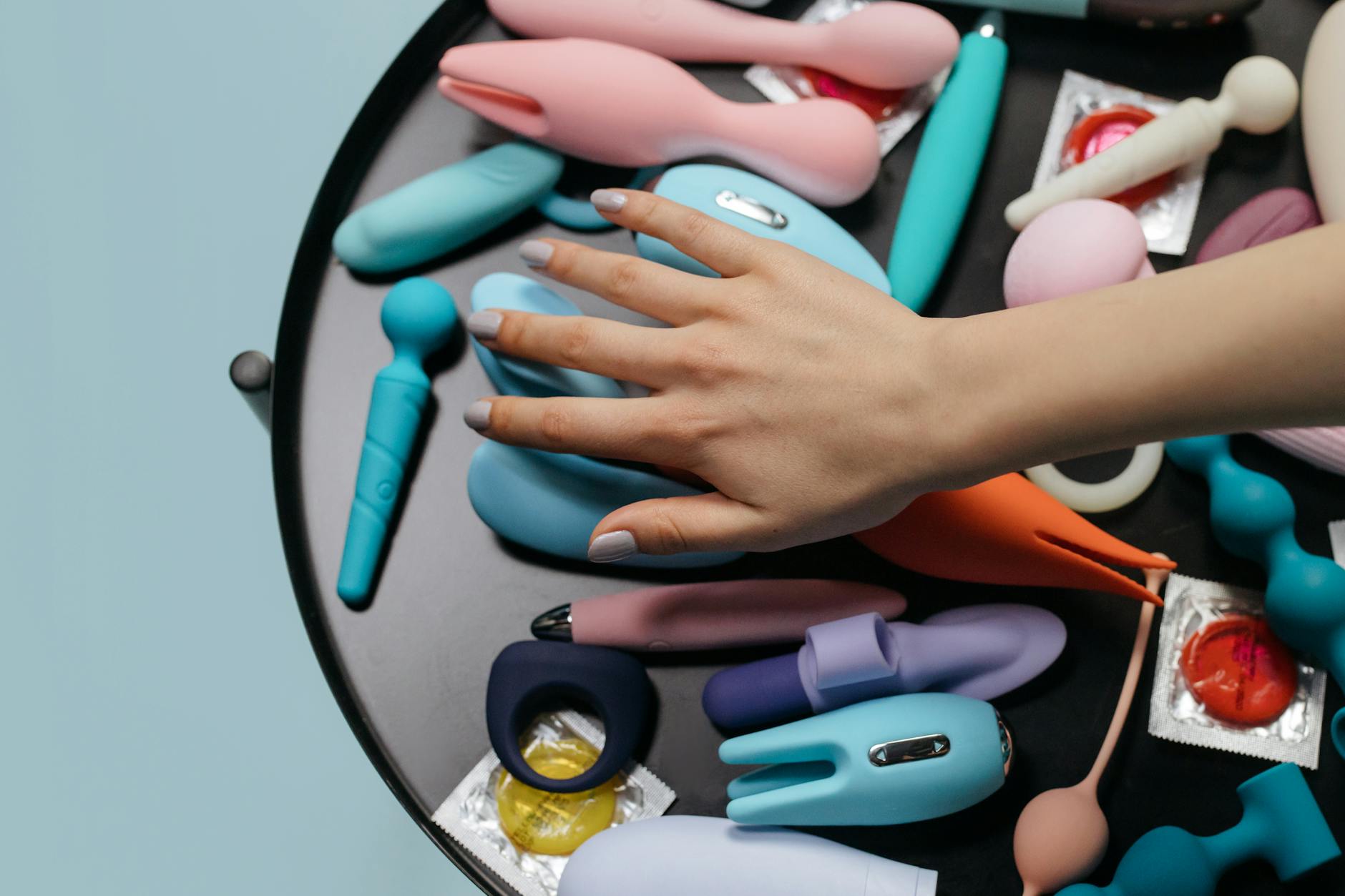 Hands reaching toward a collection of colorful vibrators and adult toys arranged on a table.